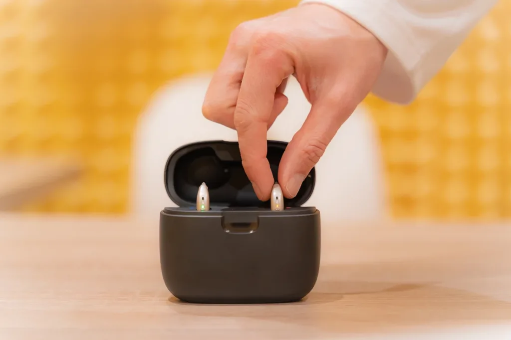 A hand of a hearing health care specialist placing rechargeable hearing aids into a stylish hearing aid charging case to charge the battery.